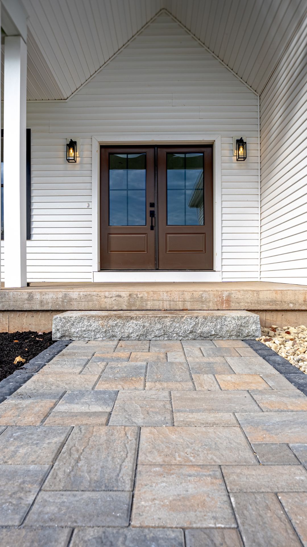 Front porch of a white house featuring double brown doors, a stone step, and a newly installed paver walkway with decorative landscaping and river rocks.